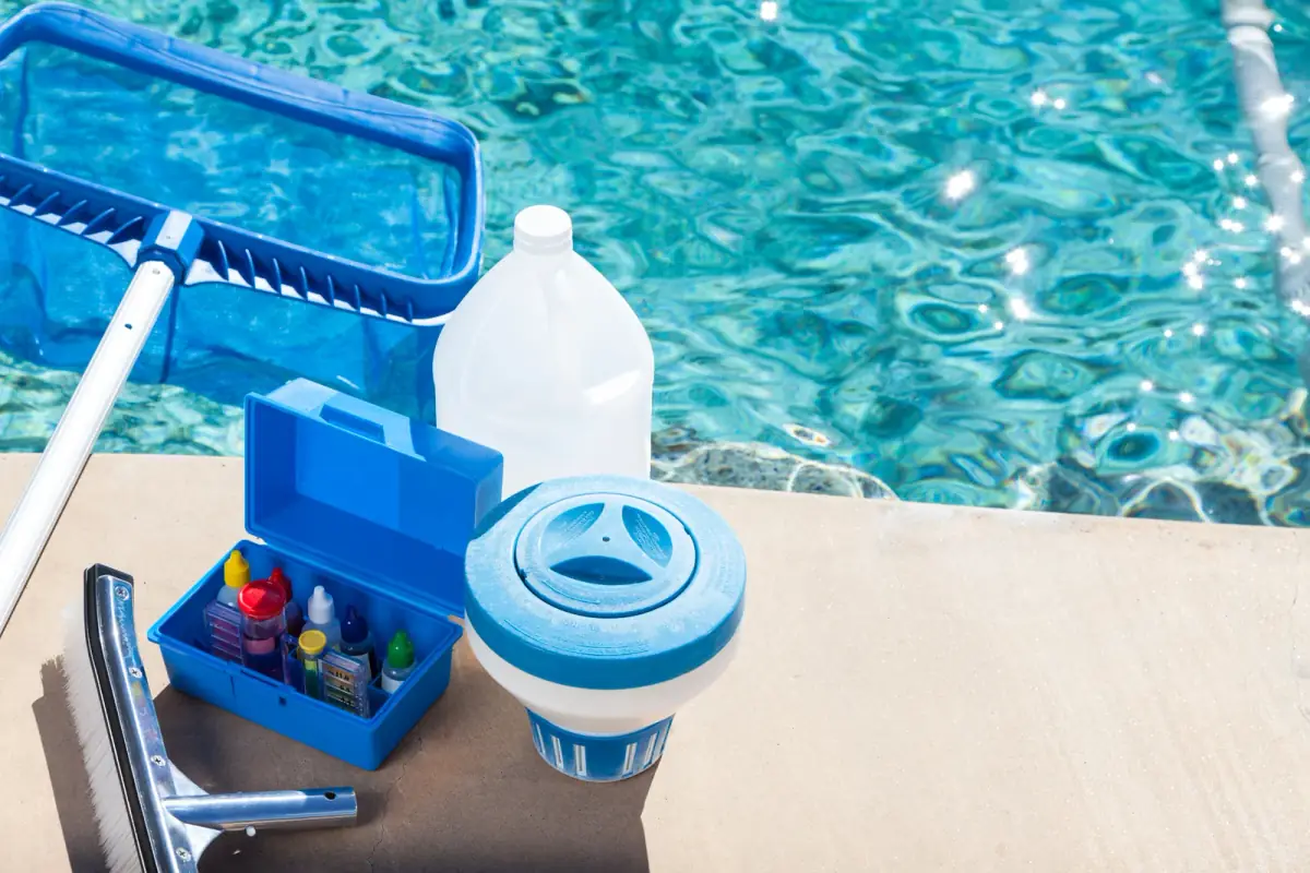 Pool chemicals, testing kit, skimmer net, and supplies arranged on a pool deck